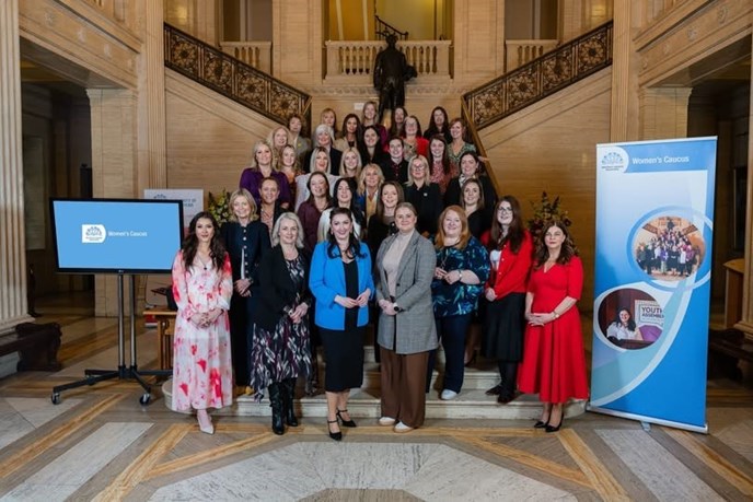 The photograph features 32 of the 36 current female MLAs with the Assembly Clerk/Chief Executive Lesley Hogg taken on the Great Hall staircase in Parliament Buildings. The MLAs are featured in rows. To the left of the image is a TV screen with the Assembly Women’s Caucus logo on it in blue and white and the words Assembly Women’s Caucus. To the right is a pop-up stand which has the Assembly blue flax logo in the top left and the words Assembly Women’s Caucus, swathes of dark and pale blue and two pictures in circles, the upper picture features all female MLAs taken on the Great Hall staircase in 2024, the bottom picture features a smiling Claire Sudden MLA, Chair of the Assembly Women’s Caucus Steering Group. At the top left and right of the picture you can see the bronze balustrades of the staircase and the statue of Lord Craigavon at the top of the stairs. You can also see the Great Hall marble to the right, left and in the background of the photograph.    , Picture, Picture